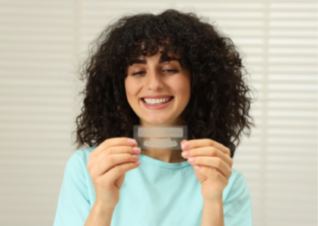 Woman holding teeth whitening strips and smiling, illustrating the use and effectiveness of whitening strips for removing surface stains and improving tooth brightness at home.